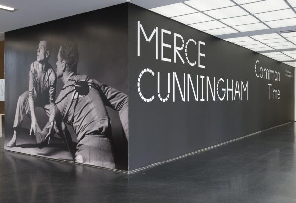 A black-walled corner in a room features a floor-to-ceiling gray-scale image of a crouching man seen from the front and the back on the left wall, and large white letters on the adjacent black wall that read: 
