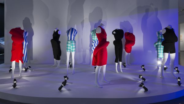 Ten headless mannequins dressed in clothing with unusual bulges are posed on a white platform and dramatically lit by spotlights on the ground.