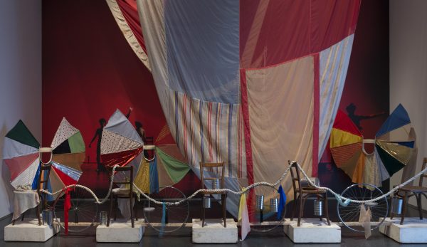 A thick white rope drapes across a row of chairs sitting on white blocks interspersed with silver bicycle tires. Behind them, a multicolor quilted fabric hangs from the ceiling in front of a red wall, and three smaller quilted fabrics fan out in imperfect circles.
