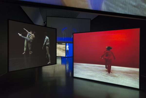 Large projection screens hung at various heights and angles fill the dimly lit gallery. Videos projected on the screens show dancers in different performances and configurations.