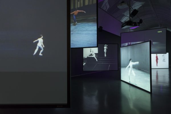 A large room is filled with projection screens hung at different heights and angles. The screens depicts dancers performing choreography.