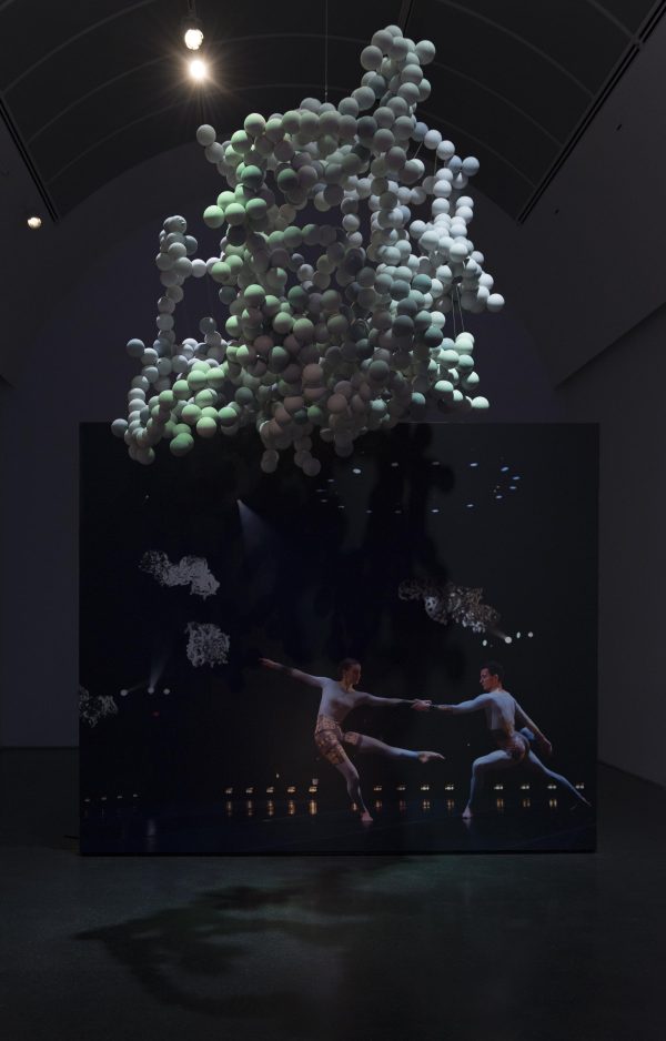 In a dark, dramatically lit space, a large amalgam of white balls hangs from the ceiling while a photo of two female dancers in unitards is visible on the wall.