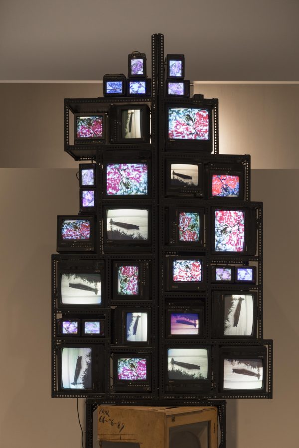Cathode ray televisions of medium to small size are stacked on top one another at varying orientations and held together by perforated metal brackets. Two different images are repeated on the different televisions: a black-and-white video of a barge on a lake and a color drawing of a flower on a patterned background.