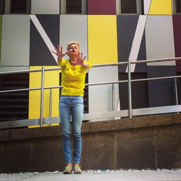 A woman poses with their hands straight out in front of a modern wall with vertical rectangular siding in greys and yellows