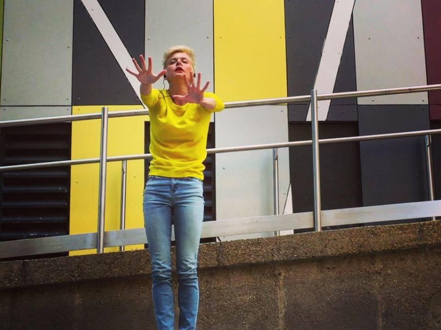 A woman poses with their hands straight out in front of a modern wall with vertical rectangular siding in greys and yellows