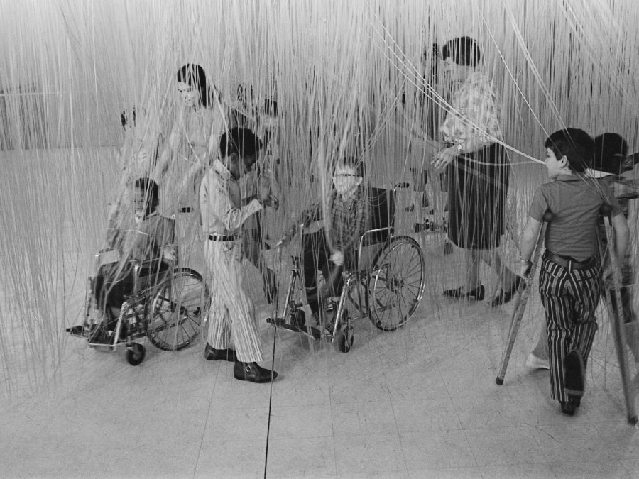 A group of children, some pushed in wheelchairs or using crutches, touches strands of an artwork hanging from the ceiling.