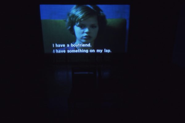 A dark room with a large screen displaying a still of a child cast in a dark blue light and the captions I have a boyfriend. I have something on my lap