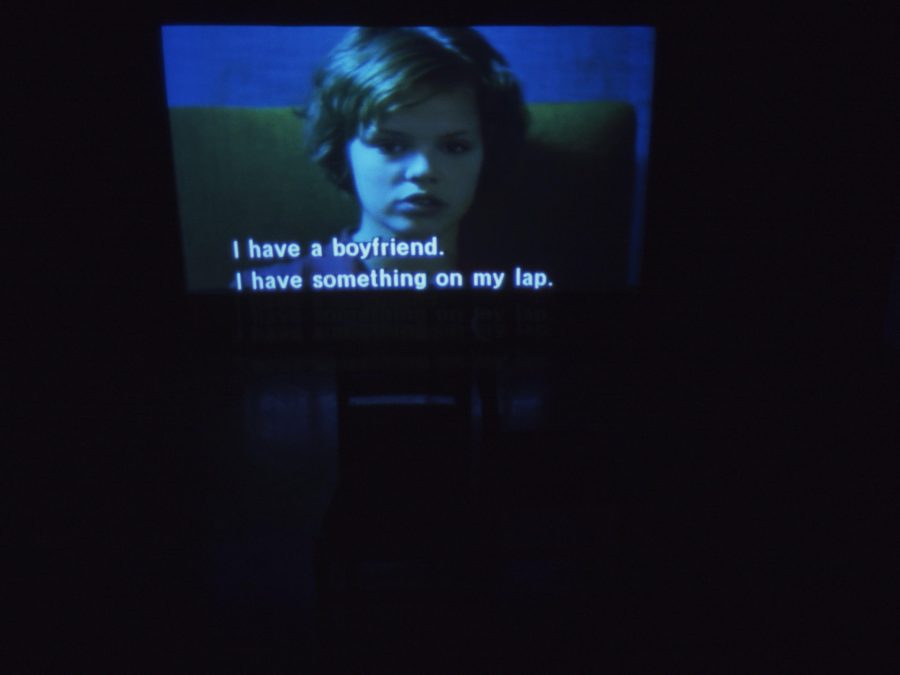A dark room with a large screen displaying a still of a child cast in a dark blue light and the captions I have a boyfriend. I have something on my lap
