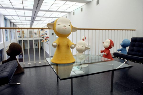 A large figurine in a painted yellow robe with eyes closed and arms out as if presenting something on a table near other similar figures of other colors including black, white, red, and blue