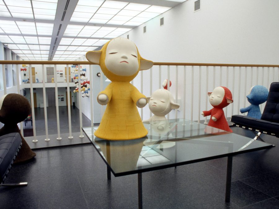 A large figurine in a painted yellow robe with eyes closed and arms out as if presenting something on a table near other similar figures of other colors including black, white, red, and blue