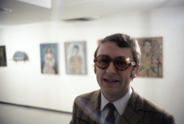 Jan van der Marck smiles, wearing dark tinted, round-framed glasses and a brown tweed sport jacket, with a white shirt and tie. Behind him are artworks hung on a white gallery wall.
