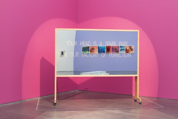 A mobile bulletin board that has been turned into a mirror that reflects a gallery with images on the walls around it. The mirror has etched into it the words: Your head is a spare part in our factory of perfection.