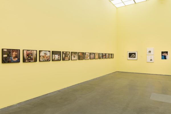 Two walls of a sherbet yellow gallery space with a line of photographs hanging on the left wall; three artworks hang on the right wall.