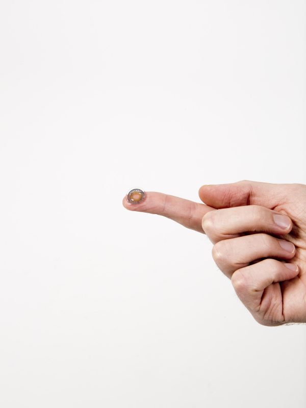 A contact lens rests on the outstretched pointer finger of a loosely clenched, light-skinned right hand.