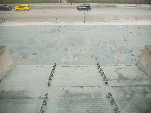 A stairway is shown from above, looking down on the plaza below where bright hazard cones dot the gray pavement tiles.