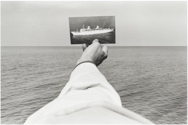 A black and white photo of a light skinned right arm wearing a white long-sleeved shirt holds a picture of a boat out over a body of water