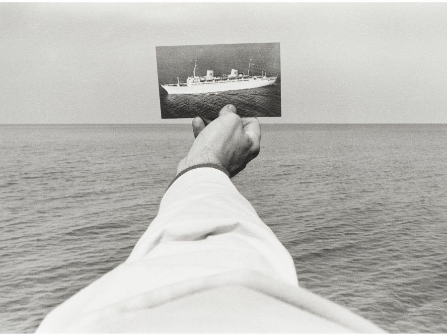 A black and white photo of a light skinned right arm wearing a white long-sleeved shirt holds a picture of a boat out over a body of water