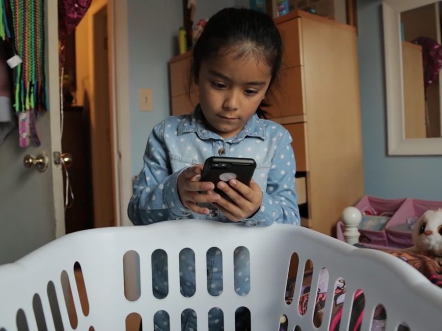 A child looks at a phone from behind a white laundry basket
