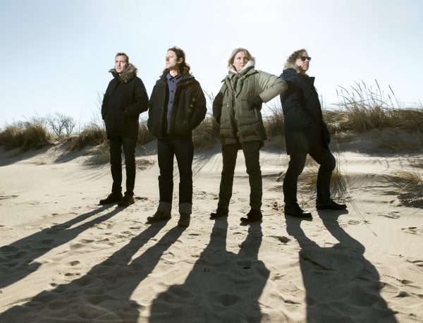 Three men and one woman stand on a beach on a sunny day wearing heavy parkas.
