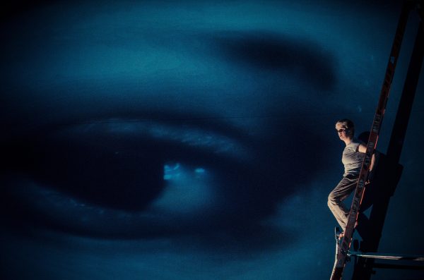 A person stands on ladder facing out against a large screen that depicts a blue closeup image of an eye