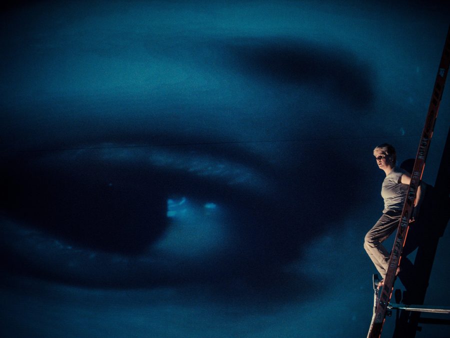 A person stands on ladder facing out against a large screen that depicts a blue closeup image of an eye