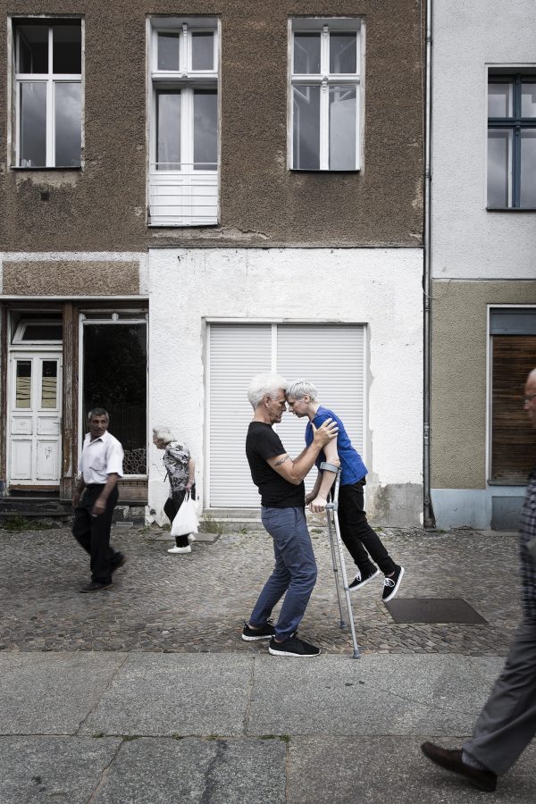 A man and woman lean on one another, their foreheads touching. The woman is held aloft by two long crutches she holds and her shoulders are steadied by the man.