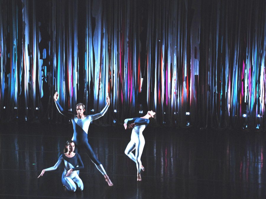 Three dancers on a stage with a shimmery curtain lit up in blues and purples behind them
