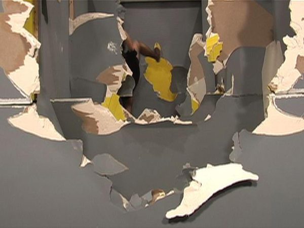 Layered gray walls are seen head on, receding into the middle distance. Each wall has a rough hole through it, forming a tunnel through which successive walls are visible. The broken edges of each hole are cream, brown, and mustard. At the end of the tunnel, a person's blurry leg wearing a yellow shoe kicks upward.