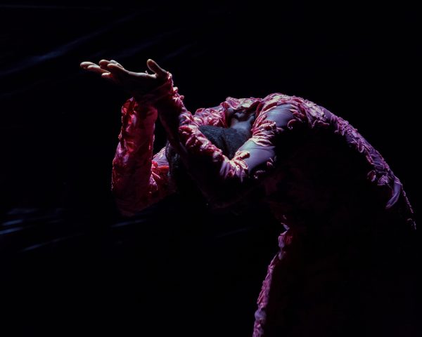 A lit figure against a dark background wears an embroidered costume and bends forward from the waist, arms raised and palms turned upward, and head faced downward, hidden from view.