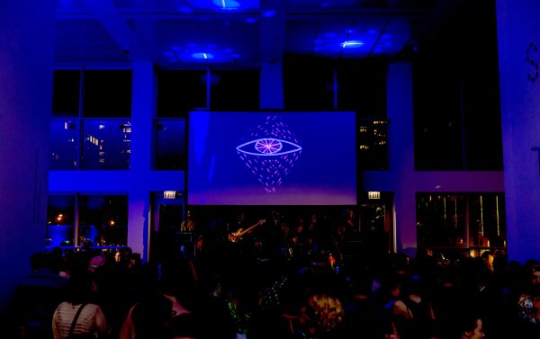 A band performs on stage under dim blue lighting, with a large screen displaying a logo of an eye with red lines forming triangles above and below it. Audience members are visible in the foreground enjoying the concert.