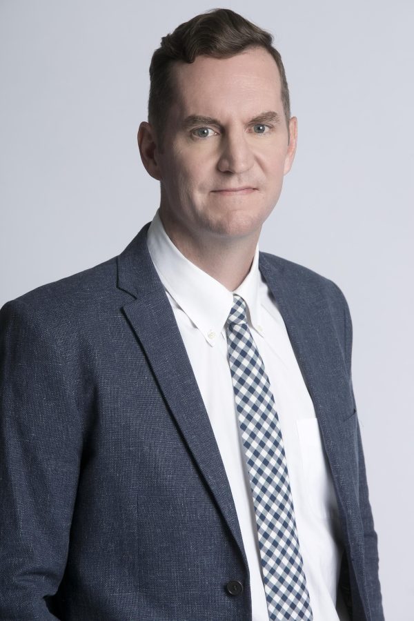 A light-skinned man wearing a blue suit jacket, white shirt, and a blue-and-white checkered tie looks directly at the viewer.