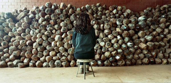A person sits on a stool facing a wall that is piled up with rocks