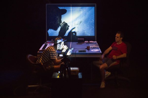 Two men sit in a dark room at a ping-pong table. One drinks from a glass and holds a lit cigarette; he is illuminated by a narrow spotlight that casts a shadow onto the vertical rear of the table.