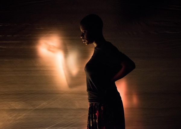 A young black woman in a dark, loose shirt and skirt stands in profile, her hands resting on the small of her back. Behind her we see the hazy glowing figure of upraised arms.