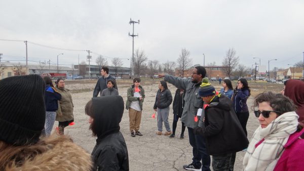 A group of people stand around an empty lot on a cold gray day. A figure in the center points to the left and several people look in that direction.
