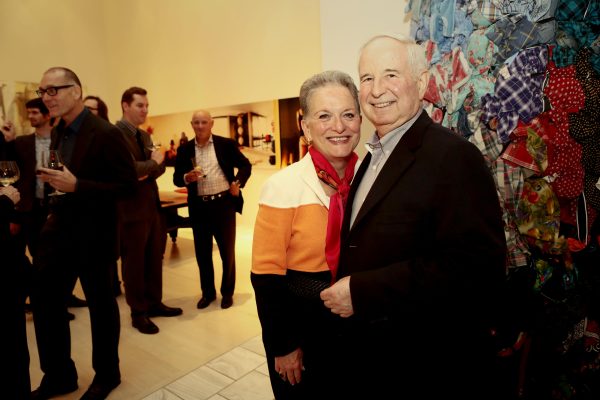 Two people with pale pink skin pose in a gallery among art and other people