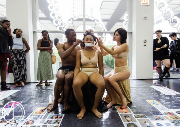 A female with tan skin wearing just a bra and underwear has tape across her mouth with illegible writing on it. Two people also in just underwear, one male presenting, the other female-presenting touch her hair as a crowd watching on.