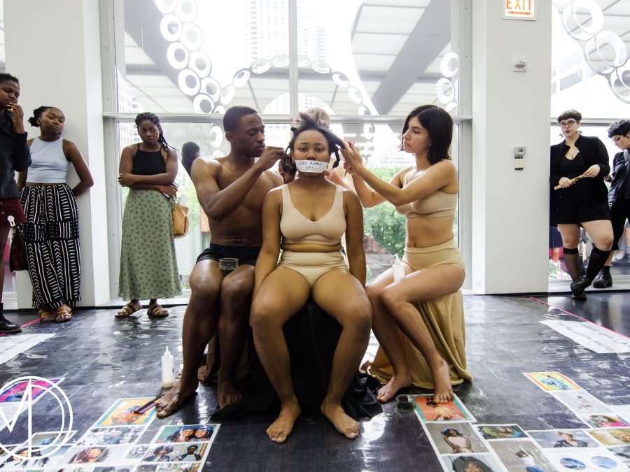 A female with tan skin wearing just a bra and underwear has tape across her mouth with illegible writing on it. Two people also in just underwear, one male presenting, the other female-presenting touch her hair as a crowd watching on.