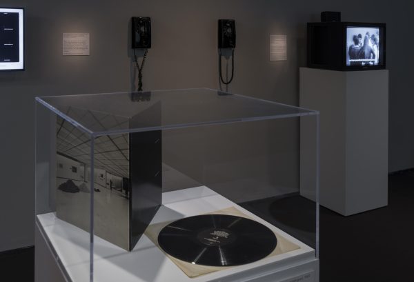 photograph of vinyl record in a display case, wall telephones and a tube television in gallery setting