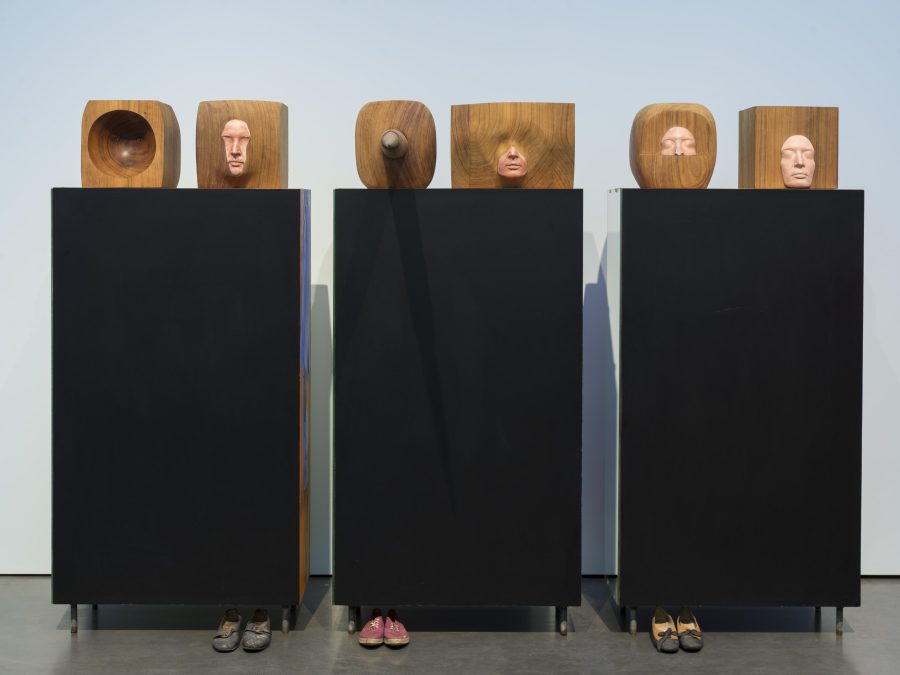 Three black boxes each with two heads carved out of brown cubes in various ways. Shoes lay at the bottom of a few of the boxes.