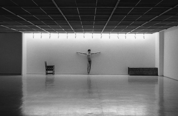 A nude person in a crucifixion pose stands at the back of a spacious, minimalist gallery with bright overhead lights, a lone chair to their left, and a bench to their right.