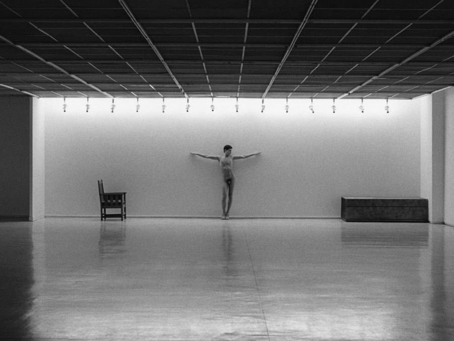 A nude person in a crucifixion pose stands at the back of a spacious, minimalist gallery with bright overhead lights, a lone chair to their left, and a bench to their right.