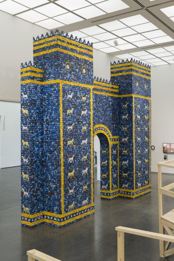 An architectural arched gateway is shown, installed within a modern gallery space. The arch is made from bricks in a range of different blues with golden yellow accents and decorated with stylized animals.