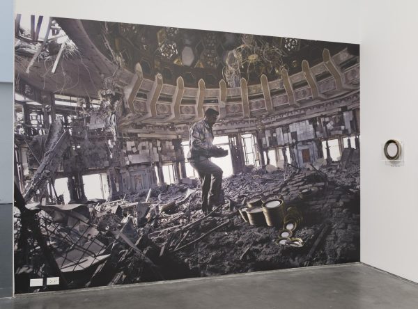 A large screen displaying an image of a person standing in the middle of a pile of rubble in what was an ornate, domed building