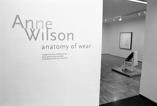 A wall with text reading Anne Wilson, in large font, anatomy of wear, in smaller font, and then illegible font below that opens to a room beyond with a chair on a platform and a painting on the wall behind it.