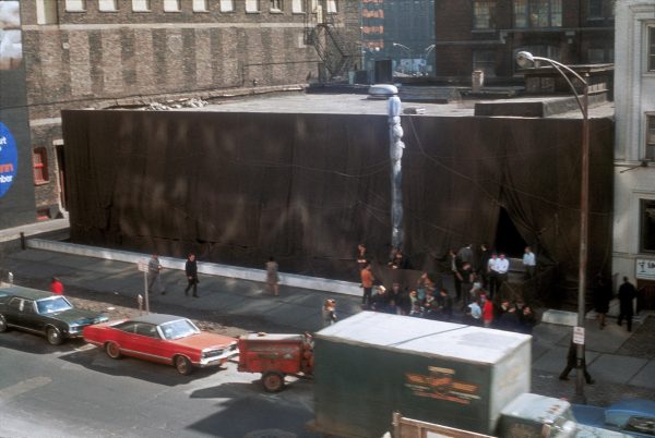 A view of a two-story building that has been wrapped in brown fabric as viewed from higher up. There is a bustle of cars and people around it.