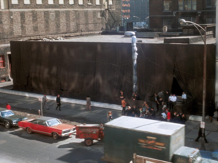 A view of a two-story building that has been wrapped in brown fabric as viewed from higher up. There is a bustle of cars and people around it.