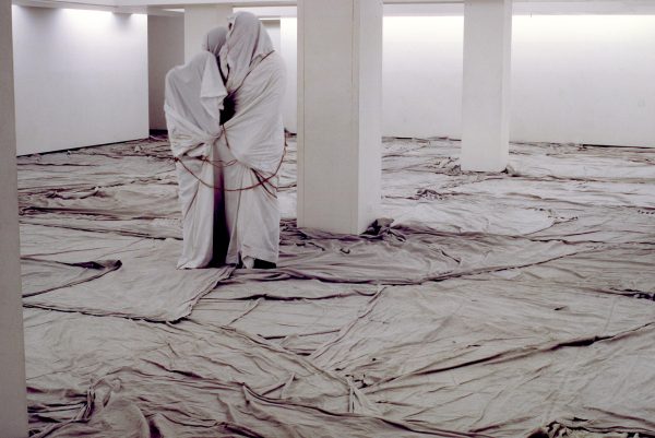 Two people wrapped in canvas and ropes stand close together in a white, columned room with more fabric covering the floor