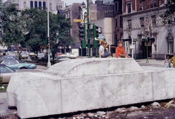 Color photograph documenting two kids sitting on a full-sized concrete car in a bustling city scene, with traffic lights and classical architecture in the background.