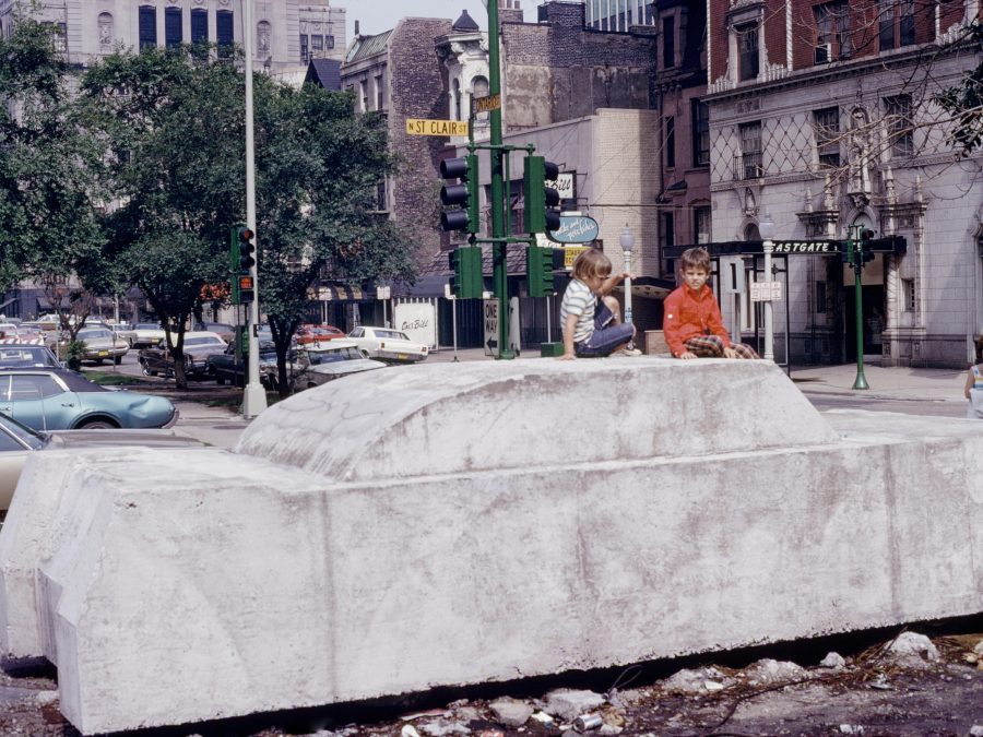 Color photograph documenting two kids sitting on a full-sized concrete car in a bustling city scene, with traffic lights and classical architecture in the background.